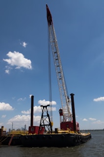 A large barge with heavy machinery, including a tall crane, is floating on a body of water. The crane is painted in red and white, towering above the vessel with a clear blue sky and a few scattered clouds in the background. There are visible red and black structures on the barge, and the water reflects a calm setting.