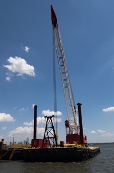 A large barge with heavy machinery, including a tall crane, is floating on a body of water. The crane is painted in red and white, towering above the vessel with a clear blue sky and a few scattered clouds in the background. There are visible red and black structures on the barge, and the water reflects a calm setting.