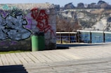 A graffiti-covered stone wall stands near a wooden boardwalk with a green trash can placed in front. The graffiti includes abstract shapes and a red drawing of a face. In the background, there is a view of a rocky coastline and some trees under a clear blue sky.
