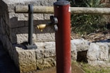 A red post-style water pump with a brass spout is positioned in front of a stone wall. A wooden beam supported by iron brackets is situated behind the pump. Vegetation is visible in the background.