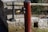 A red post-style water pump with a brass spout is positioned in front of a stone wall. A wooden beam supported by iron brackets is situated behind the pump. Vegetation is visible in the background.