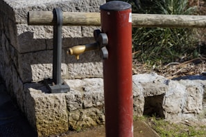 A red post-style water pump with a brass spout is positioned in front of a stone wall. A wooden beam supported by iron brackets is situated behind the pump. Vegetation is visible in the background.