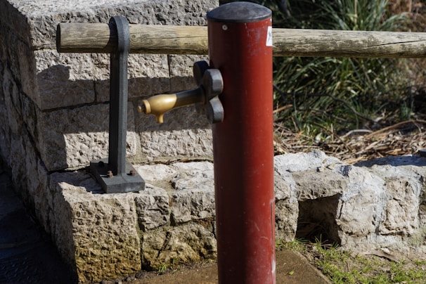 A red post-style water pump with a brass spout is positioned in front of a stone wall. A wooden beam supported by iron brackets is situated behind the pump. Vegetation is visible in the background.