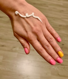 A hand with neatly manicured nails, painted in pink and one in yellow. There is a squiggle of cream or lotion on the back of the hand. The background is a wooden surface.
