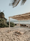 A tranquil beach setting featuring a thatched canopy providing shade over striped cushions on the sand. Palm trees sway gently in the breeze and chairs are arranged near the shoreline with the ocean visible in the background.
