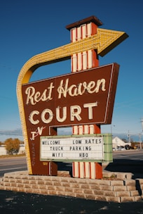 A vintage roadside motel sign stands prominently, featuring bold retro typography and an arrow design pointing toward the motel. The sign reads 'Rest Haven Court' and advertises amenities like low rates, truck parking, Wi-Fi, and HBO. The background includes a clear blue sky and a suburban road setting with some trees and buildings visible.