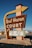A vintage roadside motel sign stands prominently, featuring bold retro typography and an arrow design pointing toward the motel. The sign reads 'Rest Haven Court' and advertises amenities like low rates, truck parking, Wi-Fi, and HBO. The background includes a clear blue sky and a suburban road setting with some trees and buildings visible.