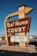 A vintage roadside motel sign stands prominently, featuring bold retro typography and an arrow design pointing toward the motel. The sign reads 'Rest Haven Court' and advertises amenities like low rates, truck parking, Wi-Fi, and HBO. The background includes a clear blue sky and a suburban road setting with some trees and buildings visible.