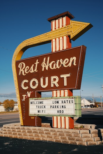 A vintage roadside motel sign stands prominently, featuring bold retro typography and an arrow design pointing toward the motel. The sign reads 'Rest Haven Court' and advertises amenities like low rates, truck parking, Wi-Fi, and HBO. The background includes a clear blue sky and a suburban road setting with some trees and buildings visible.
