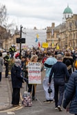 A large crowd gathered on a city street, holding signs and banners. One prominent sign in the foreground reads 'SAY NO TO 15-MIN GHETTOS.' The participants appear to be engaged in a protest against an urban planning concept. Several people are wearing casual clothing, and police officers are visible to the left, managing the event. The buildings in the background have historic architectural features.