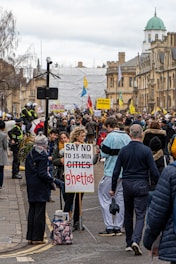 A large crowd gathered on a city street, holding signs and banners. One prominent sign in the foreground reads 'SAY NO TO 15-MIN GHETTOS.' The participants appear to be engaged in a protest against an urban planning concept. Several people are wearing casual clothing, and police officers are visible to the left, managing the event. The buildings in the background have historic architectural features.