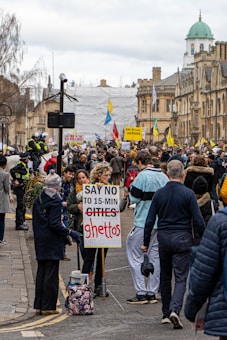 A large crowd gathered on a city street, holding signs and banners. One prominent sign in the foreground reads 'SAY NO TO 15-MIN GHETTOS.' The participants appear to be engaged in a protest against an urban planning concept. Several people are wearing casual clothing, and police officers are visible to the left, managing the event. The buildings in the background have historic architectural features.