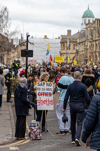 A large crowd gathered on a city street, holding signs and banners. One prominent sign in the foreground reads 'SAY NO TO 15-MIN GHETTOS.' The participants appear to be engaged in a protest against an urban planning concept. Several people are wearing casual clothing, and police officers are visible to the left, managing the event. The buildings in the background have historic architectural features.