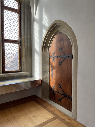 An interior setting featuring a tall, arched wooden door with intricate black metal hinges and handles. The door is set within a stone frame against a plain white wall. To the left of the door, a window with diamond-patterned panes allows natural light to enter, casting shadows on the wall. The floor is made of polished wood, adding warmth to the space.