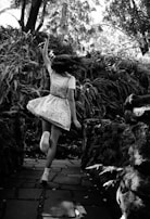 A girl in a dress is dancing joyfully on a stone pathway surrounded by dense foliage and tall trees. Her hair is in motion, suggesting movement and freedom.