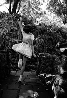 A girl in a dress is dancing joyfully on a stone pathway surrounded by dense foliage and tall trees. Her hair is in motion, suggesting movement and freedom.