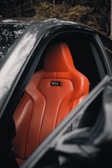 A sleek orange leather car seat is prominently featured inside a luxury vehicle. The design is ergonomic with visible stitching and contouring for comfort. The surrounding area is composed of a dark interior and part of the car's door frame.