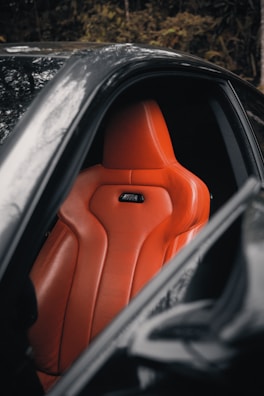 A sleek orange leather car seat is prominently featured inside a luxury vehicle. The design is ergonomic with visible stitching and contouring for comfort. The surrounding area is composed of a dark interior and part of the car's door frame.