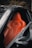 A sleek orange leather car seat is prominently featured inside a luxury vehicle. The design is ergonomic with visible stitching and contouring for comfort. The surrounding area is composed of a dark interior and part of the car's door frame.