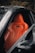 A sleek orange leather car seat is prominently featured inside a luxury vehicle. The design is ergonomic with visible stitching and contouring for comfort. The surrounding area is composed of a dark interior and part of the car's door frame.