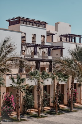 A modern building features a series of terraces with decorative pergolas on the upper floors. The facade is complemented by tall palm trees and vibrant flowering shrubs in the foreground, creating a harmonious blend of architecture and nature. The sky is clear and blue.