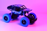 A toy off-road vehicle with blue tires and black body is set against a vibrant pink and purple background. The vehicle's frame is detailed with a pattern and the words 'Super Off-Road' on the side, adding to its rugged appearance.