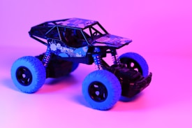 A toy off-road vehicle with blue tires and black body is set against a vibrant pink and purple background. The vehicle's frame is detailed with a pattern and the words 'Super Off-Road' on the side, adding to its rugged appearance.