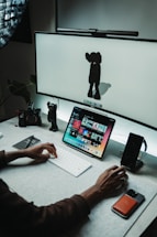 A workspace with a computer setup featuring a large screen displaying a minimalist character and a tablet showing a variety of colorful images. A person's hand is actively using a keyboard in front of the devices. There are a few desk accessories, including a camera and a smartphone, along with a small character figurine near the edge.