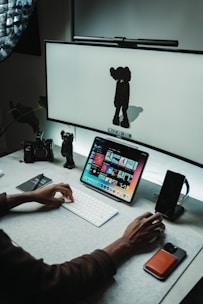 A workspace with a computer setup featuring a large screen displaying a minimalist character and a tablet showing a variety of colorful images. A person's hand is actively using a keyboard in front of the devices. There are a few desk accessories, including a camera and a smartphone, along with a small character figurine near the edge.
