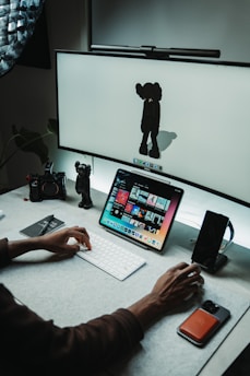 A workspace with a computer setup featuring a large screen displaying a minimalist character and a tablet showing a variety of colorful images. A person's hand is actively using a keyboard in front of the devices. There are a few desk accessories, including a camera and a smartphone, along with a small character figurine near the edge.