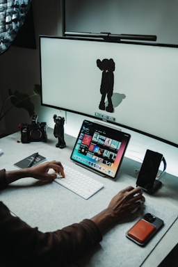 A workspace with a computer setup featuring a large screen displaying a minimalist character and a tablet showing a variety of colorful images. A person's hand is actively using a keyboard in front of the devices. There are a few desk accessories, including a camera and a smartphone, along with a small character figurine near the edge.