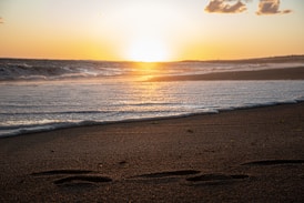 the sun is setting over the ocean with footprints in the sand