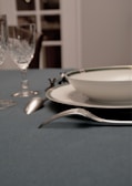 A dining table setting features a blue tablecloth, a white plate with a bordered design, a silver fork and spoon, and two crystal glasses. The background has soft focus, with visible sections of a window or door.