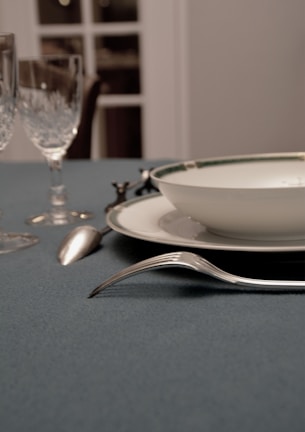 A dining table setting features a blue tablecloth, a white plate with a bordered design, a silver fork and spoon, and two crystal glasses. The background has soft focus, with visible sections of a window or door.