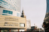A modern urban scene with a large commercial building displaying various brand logos such as Bershka, Pull&Bear, McDonald's, and Media Markt. The architecture is contemporary with glass panels and geometric shapes. A historic tower is visible in the background, presenting a contrast of styles. The image has a clear sky and a street lamp.