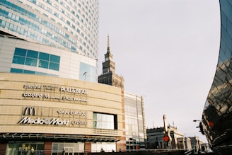 A modern urban scene with a large commercial building displaying various brand logos such as Bershka, Pull&Bear, McDonald's, and Media Markt. The architecture is contemporary with glass panels and geometric shapes. A historic tower is visible in the background, presenting a contrast of styles. The image has a clear sky and a street lamp.