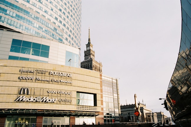A modern urban scene with a large commercial building displaying various brand logos such as Bershka, Pull&Bear, McDonald's, and Media Markt. The architecture is contemporary with glass panels and geometric shapes. A historic tower is visible in the background, presenting a contrast of styles. The image has a clear sky and a street lamp.