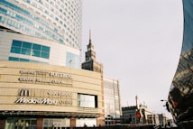 A modern urban scene with a large commercial building displaying various brand logos such as Bershka, Pull&Bear, McDonald's, and Media Markt. The architecture is contemporary with glass panels and geometric shapes. A historic tower is visible in the background, presenting a contrast of styles. The image has a clear sky and a street lamp.