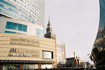 A modern urban scene with a large commercial building displaying various brand logos such as Bershka, Pull&Bear, McDonald's, and Media Markt. The architecture is contemporary with glass panels and geometric shapes. A historic tower is visible in the background, presenting a contrast of styles. The image has a clear sky and a street lamp.