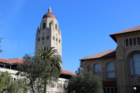 A tall tower with a red-tiled dome stands prominently against a clear blue sky. Surrounding the tower are university buildings with similar red-tiled roofs and large, arched windows. A palm tree and other greenery enhance the foreground, adding a natural element to the architectural scene.