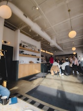 A cozy cafe interior with patrons seated around tables. The decor includes bright, overhead lighting and modern furnishings. The ceiling is high with white beams and there is a counter with wooden cabinets and a 'Complete Imported Car Service' sign. People are casually dressed, some engaged in conversation, and there is a relaxed atmosphere.