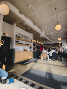 A cozy cafe interior with patrons seated around tables. The decor includes bright, overhead lighting and modern furnishings. The ceiling is high with white beams and there is a counter with wooden cabinets and a 'Complete Imported Car Service' sign. People are casually dressed, some engaged in conversation, and there is a relaxed atmosphere.
