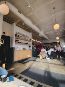 A cozy cafe interior with patrons seated around tables. The decor includes bright, overhead lighting and modern furnishings. The ceiling is high with white beams and there is a counter with wooden cabinets and a 'Complete Imported Car Service' sign. People are casually dressed, some engaged in conversation, and there is a relaxed atmosphere.