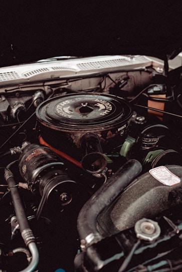 An overhead view of a vintage car's engine compartment, featuring a round air cleaner with visible branding on top, encircled by various metallic components, pipes, and wires.