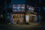 A cozy, illuminated Japanese restaurant with traditional signage and paper lanterns. The entrance features various colorful posters and a lighted menu board displaying offerings and prices. The setting is in an urban environment, captured during nighttime, giving a warm and inviting appearance.