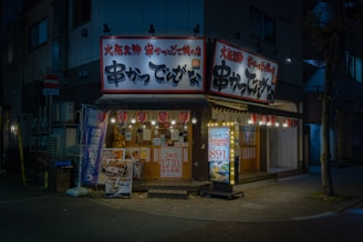 A cozy, illuminated Japanese restaurant with traditional signage and paper lanterns. The entrance features various colorful posters and a lighted menu board displaying offerings and prices. The setting is in an urban environment, captured during nighttime, giving a warm and inviting appearance.