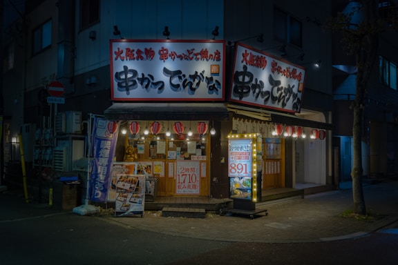 A cozy, illuminated Japanese restaurant with traditional signage and paper lanterns. The entrance features various colorful posters and a lighted menu board displaying offerings and prices. The setting is in an urban environment, captured during nighttime, giving a warm and inviting appearance.