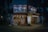 A cozy, illuminated Japanese restaurant with traditional signage and paper lanterns. The entrance features various colorful posters and a lighted menu board displaying offerings and prices. The setting is in an urban environment, captured during nighttime, giving a warm and inviting appearance.