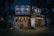 A cozy, illuminated Japanese restaurant with traditional signage and paper lanterns. The entrance features various colorful posters and a lighted menu board displaying offerings and prices. The setting is in an urban environment, captured during nighttime, giving a warm and inviting appearance.