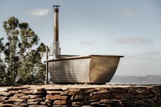 A rustic, metal-clad hot tub with a chimney sits on top of a stone wall in an outdoor setting. There is a tree in the background and a distant view of mountains under a blue sky with scattered clouds.